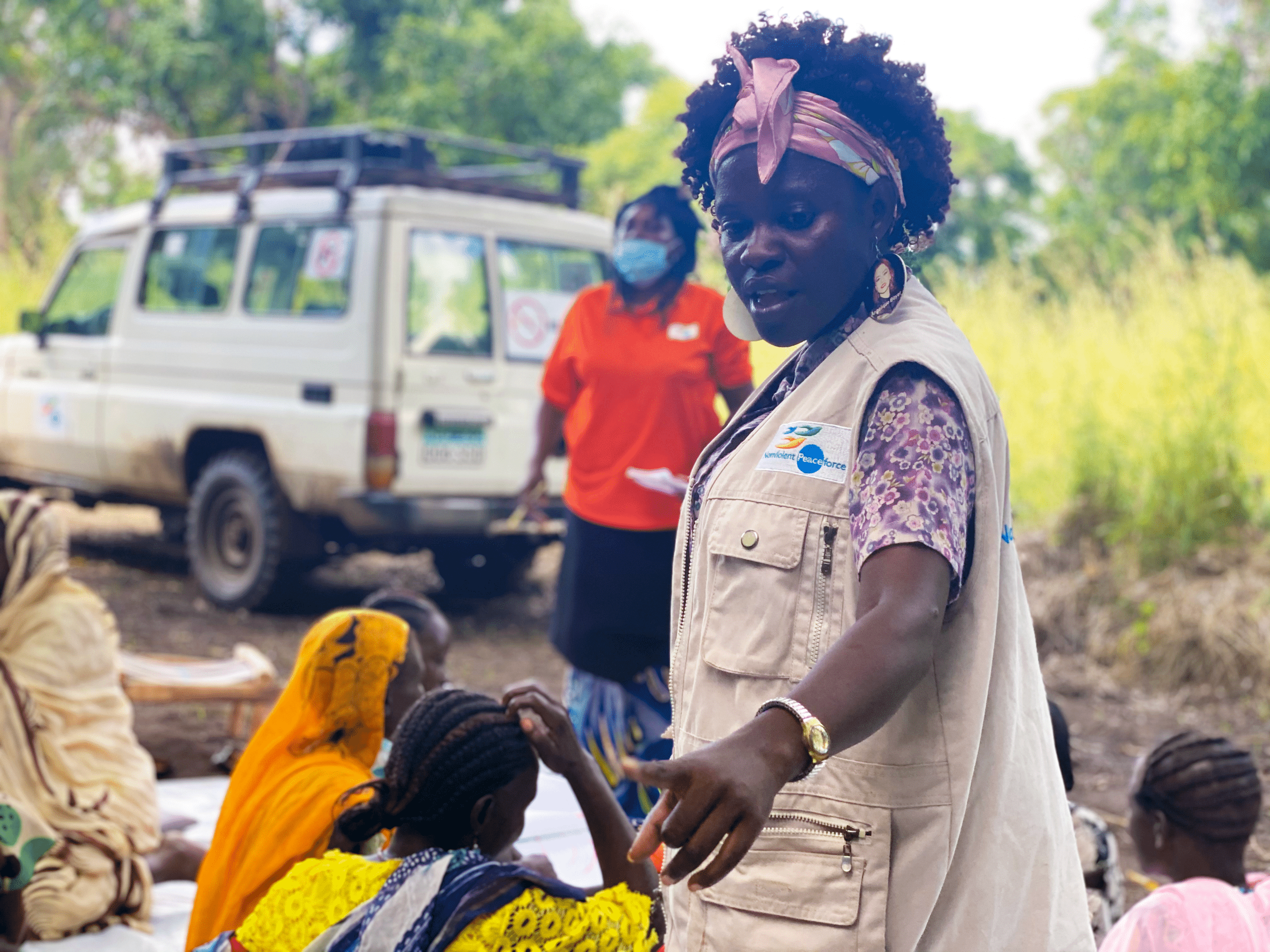 People facing two NP staff - one man in orange shirt and one woman in NP vest, in front of blackboard in South Sudan