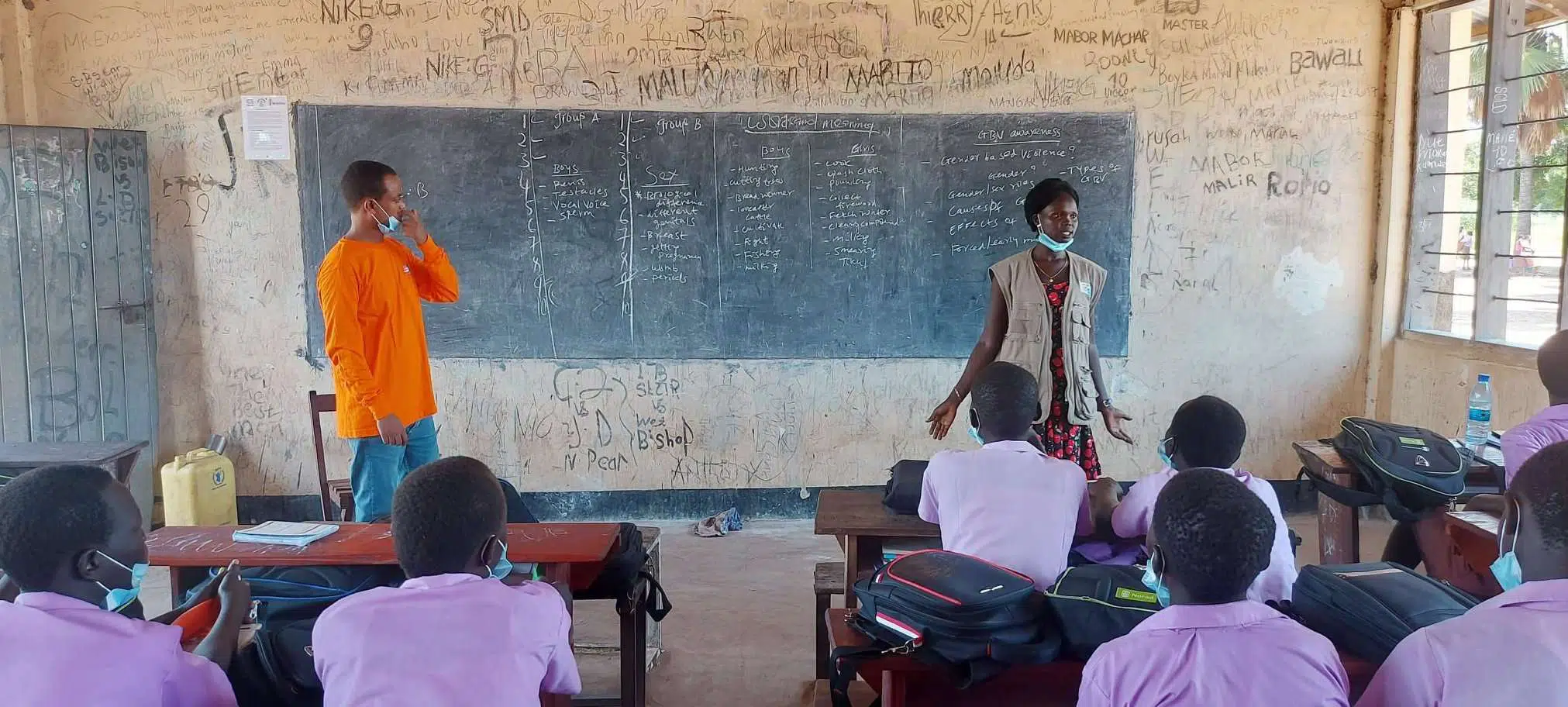 People facing two NP staff - one man in orange shirt and one woman in NP vest, in front of blackboard in South Sudan