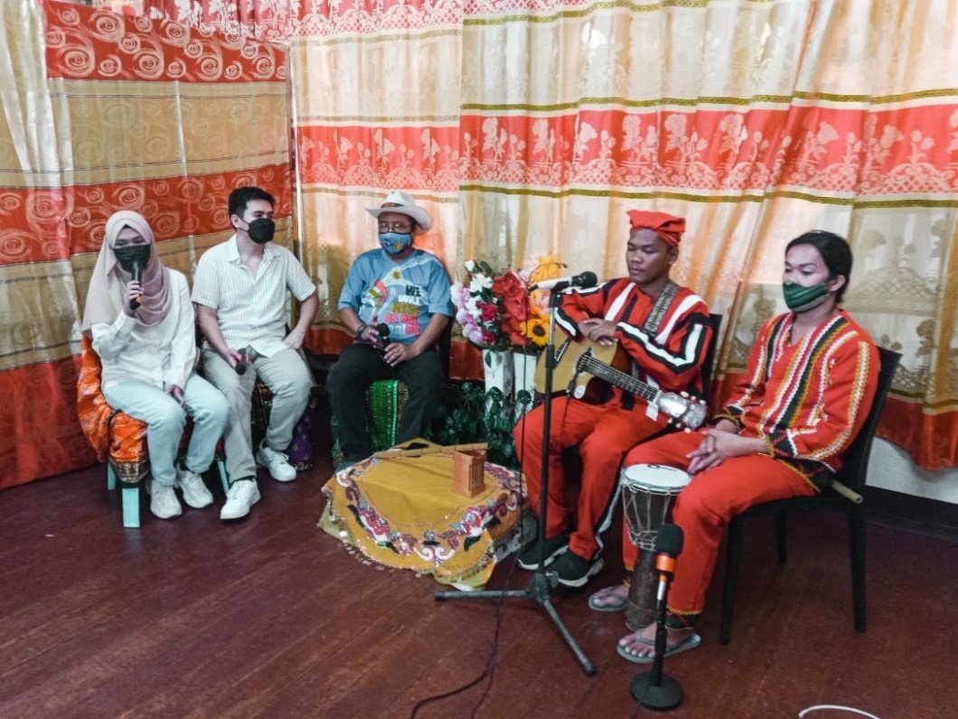 Merhanky, Neil et trois autres personnes sont assis autour du micro de diffusion. Il y a des fleurs et un fond à motifs. Les autres personnes sur la photo tiennent des instruments.