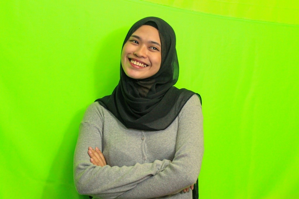 Merhanky smiles and stands against a lime green background. She wears a black headscarf and a gray shirt