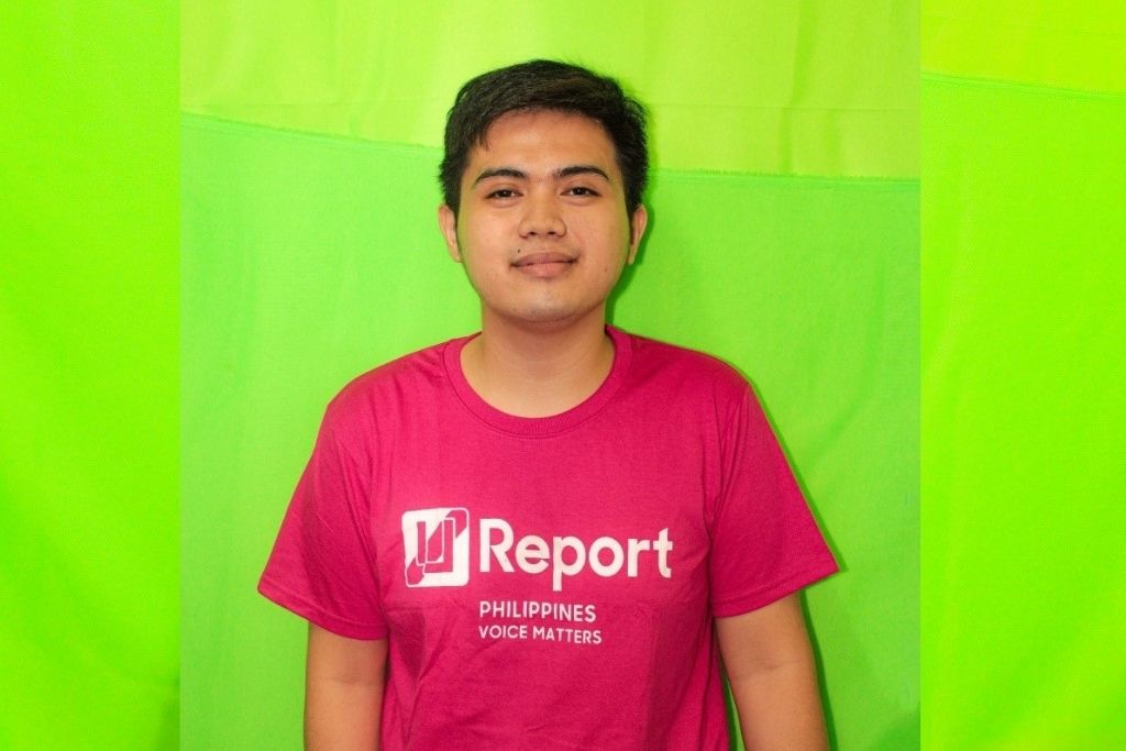 Neil smiles and stands against a lime green background. He wears a pink shirt that reads “Report. Philippines Voice Matters.”