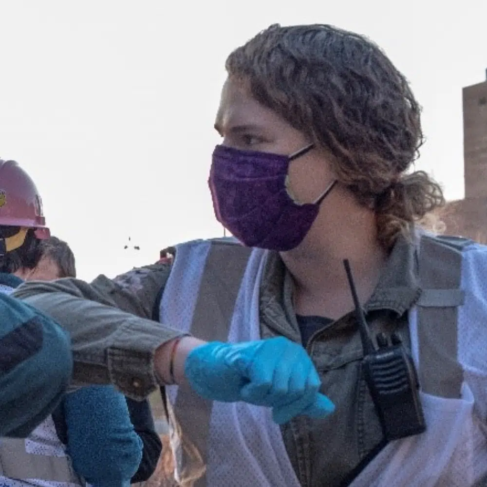 Une personne dont les cheveux sont tirés en chignon bas accueille un collègue en se cognant les coudes. Elle porte un masque, des gants, un gilet NP et un microphone.