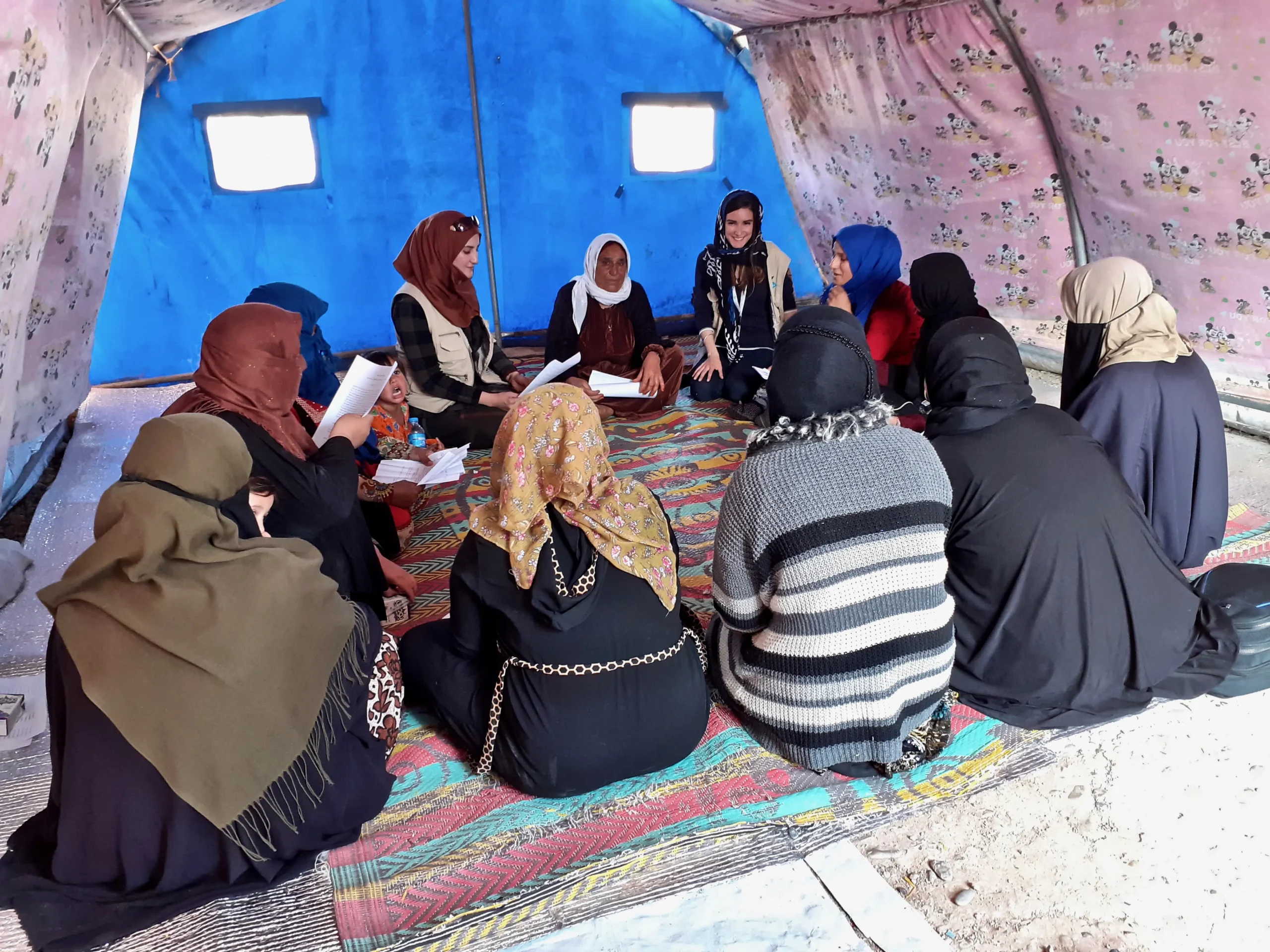 A group of women sit in a circle of the floor of a makeshift tent. They are engaged in a discussion.