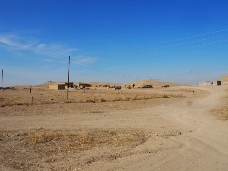 A vast, dry plain with sparse vegetation; a dirt road running through the center; utility poles on both sides of the road; an open sky above; a clear, blue day.