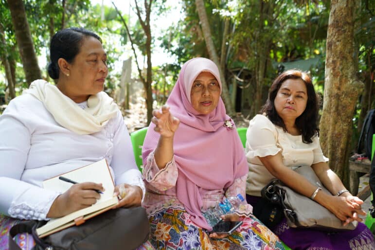 Three individuals engage in discussion outdoors: a person gestures as they speak, another person writing in a notebook, and a third person seated beside them.