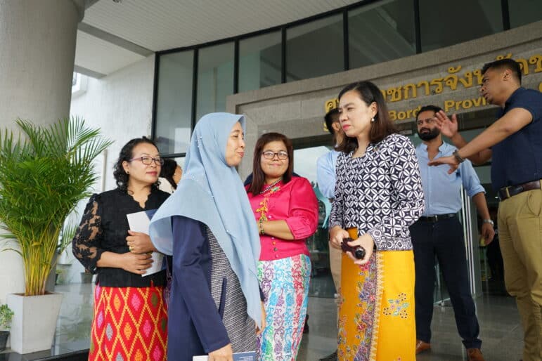 A group of people engaged in a conversation outside of a building in Thailand. One person is gesturing while standing.
