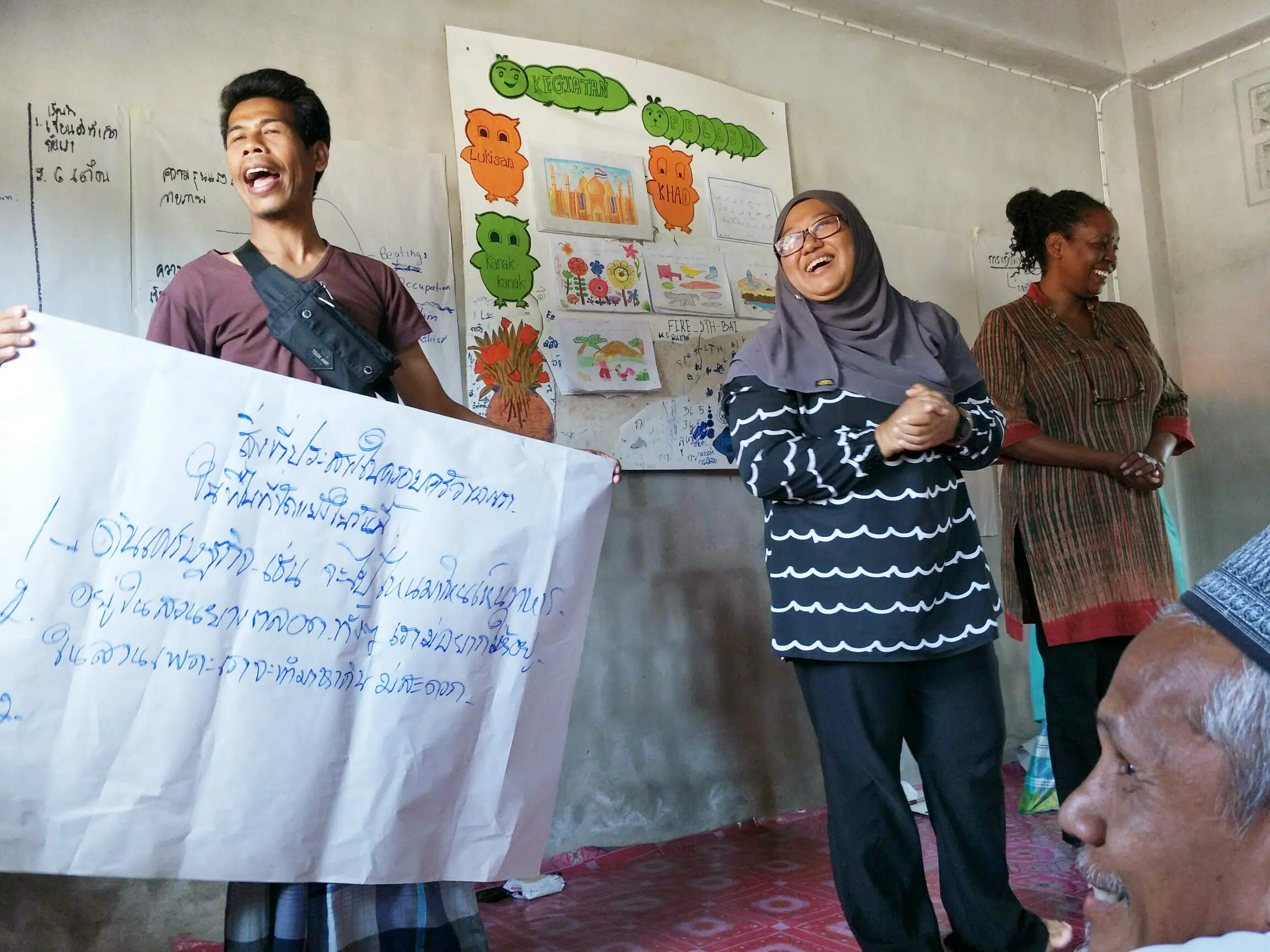 Dos personas cantando y sonriendo sosteniendo un cartel con letras tailandesas durante un entrenamiento en el sur de Tailandia. Personal de NP sonriendo junto a dos personas.