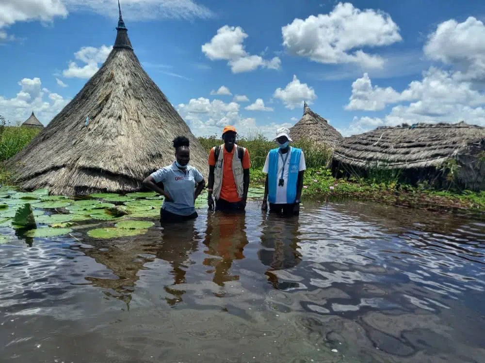 Three people, including 2 NP staff members, wade through the water in front of 4 flooded huts.