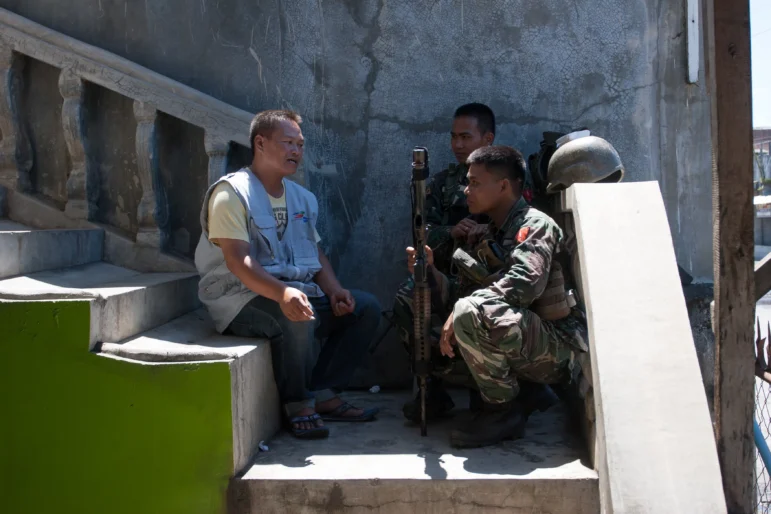 two uniformed individuals in conversation with one NP staff member. They share a cigarette and sit on the steps.