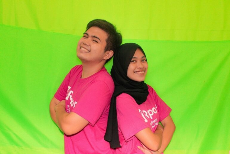 Two youth members stand back to back smiling at the camera. one man wearing a pink shirt and one woman wearing a pink shirt and black hijab. they are standing in front of a green screen.