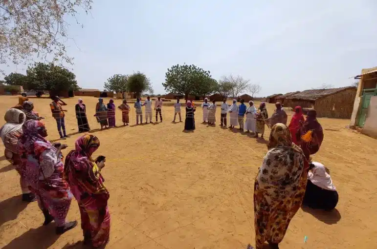 Personal de Nonviolent Peaceforce y miembros de la comunidad de Zamzam forman un círculo al aire libre. Están realizando un ejercicio de construcción de redes de paz durante una capacitación de protección comunitaria. Cada persona sostiene un hilo que se conecta formando una gran red.