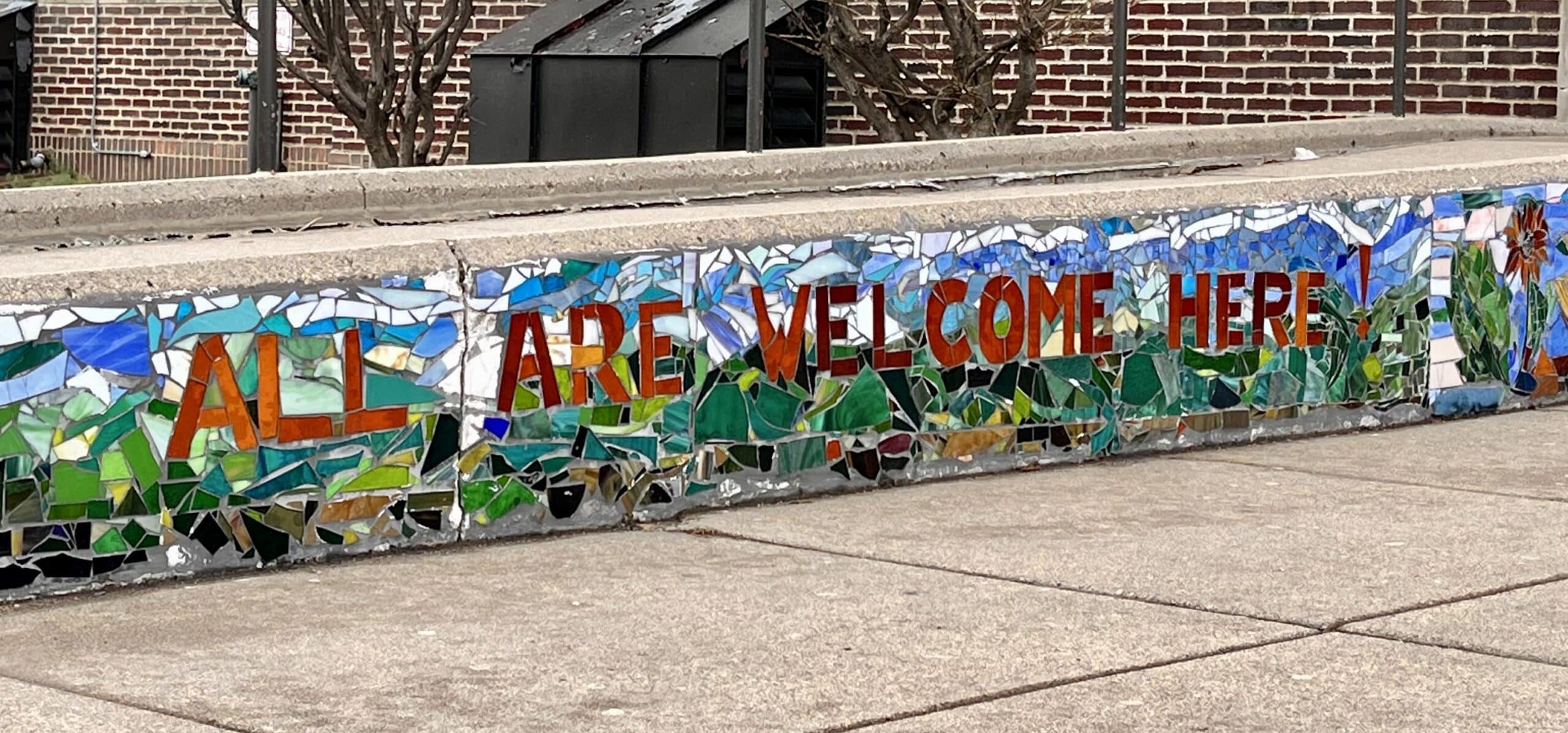 Wall mural mosaic sign reads, All are welcome here! in burnt orange. White, blue, and green mixed titles fill the background.