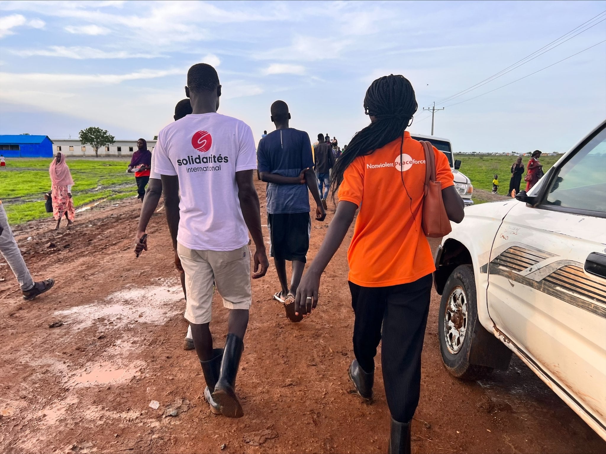 Solidarites International and Nonviolent Peaceforce staff members area walking together side-by-side. Their backs are turned to the camera as they walk along a dirt road. Solidarites uniform shirt is white and the Nonviolent Peaceforce uniform is bright orange. 