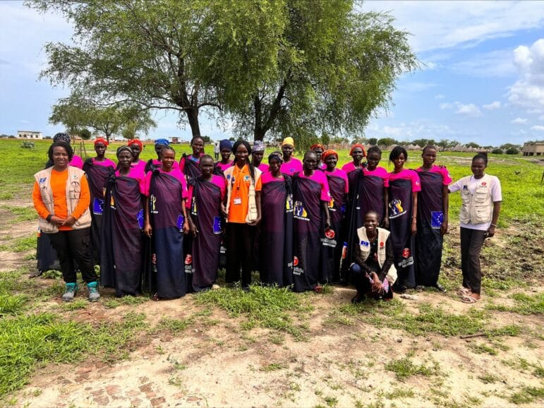 Members of the Women Civic Action Group (WCAG) are standing together outdoors with Nonviolent Peaceforce and Solidarites International staff. The WCAG members are wearing dark blue lawas and pink t-shirts. Location is Pariak, a village in Upper Nile of Melut County, South Sudan.