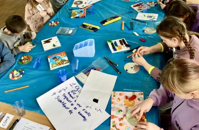A group of kids attending a workshop in Izyum Ukraine. They gather around a table, each one is focused on completing their craft project with clay, old CDs, paper, colored pencils, and more.