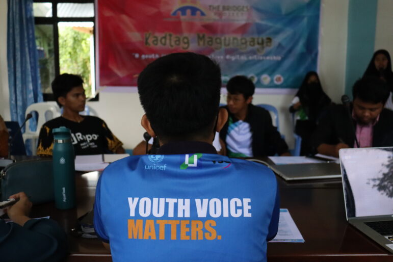 Los jóvenes que asisten al taller se sientan alrededor de una mesa larga y conversan. La camiseta de uno de ellos dice "La voz de los jóvenes importa".