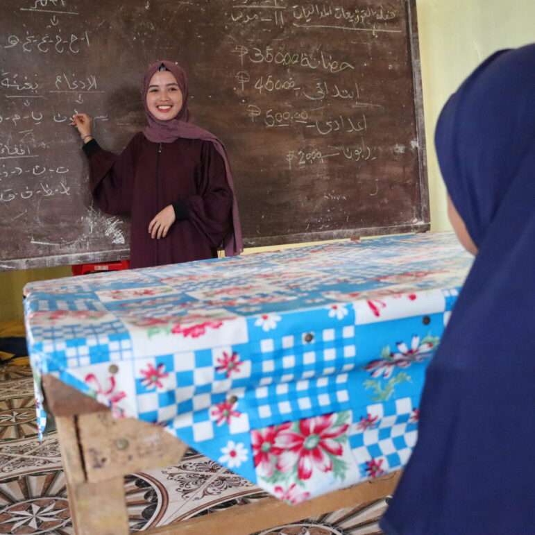 Eine philippinische Lehrerin steht vor einem Klassenzimmer und schreibt an die Tafel. Sie trägt ein lilafarbenes Kopftuch und ein pflaumenfarbenes Kleid.