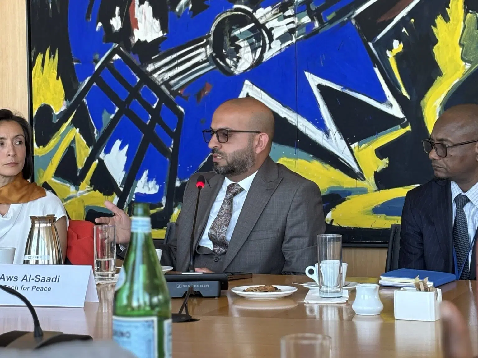 Aws A/-Saadi, a peace speaker, is seated at a conference table with a microphone positioned in front of him. He is accompanied by two other individuals seated behind him. Behind the group hangs a vibrant piece of artwork featuring bright colors, including blue, yellow, and black.