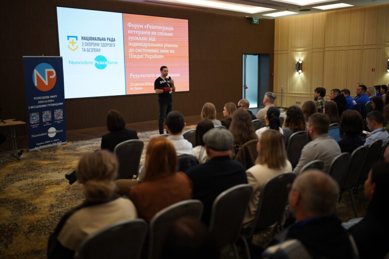 Eine Person steht mit einem Mikrofon in der Hand vor dem Publikum während einer Veranstaltung zur Wiedereingliederung von Veteranen in Odessa, Ukraine. Hinter ihr wird eine PowerPoint-Präsentation auf einer Leinwand gezeigt.
