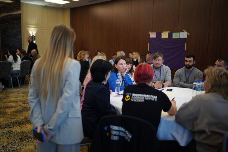 Eine Gruppe von Teilnehmern sitzt in einem Konferenzraum in Odessa um einen runden Tisch und diskutiert im Rahmen eines Veteranenforums. Ein Moderator steht in der Nähe, während die Anwesenden zuhören und ihre Ideen austauschen.