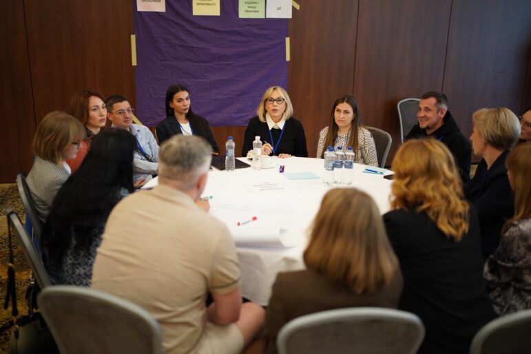 Die Teilnehmer sitzen im Kreis um einen Tisch und hören einer Frau zu, die in einer Gruppendiskussion spricht. Sie tragen Namensschilder und wirken konzentriert und aufmerksam. Die Veranstaltung findet in einem formellen Konferenzraum statt, die Workshop-Materialien liegen auf dem Tisch bereit.