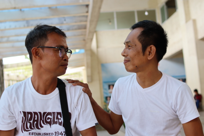 Two men in the Philippines sitting down side-by-side., faces towards each other. One man places his hand on the shoulder of the other guy in a display of forgiveness.