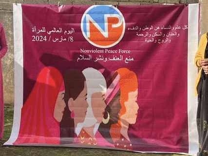 Women are standing outdoors holding purple flowers in their hands during an International Women's Day celebration. Between them is a banner with the Nonviolent Peaceforce logo. The background of the banner is a deep purple (jam colored) and features an illustration of five women. Many of the women are also wearing deep purple articles of clothing such as scarves, headscarves, and dresses. Location is: Zummar, Iraq.