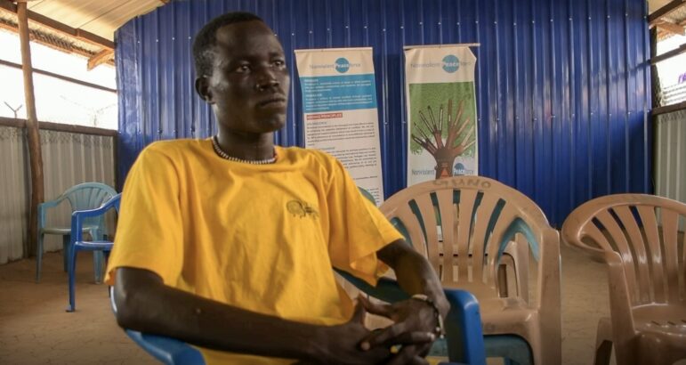A young man, age 24 sits in a chair during an interview. He's wearing a yellow shirt with a Youth Protection Team logo on it.