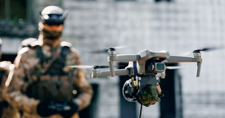 A multi-rotor drone flies in front of a soldier holding a controller.