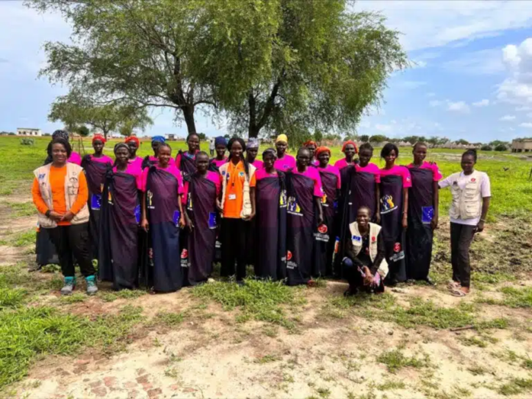 A group of women, many wearing pink t-shirts, pose for a photo outdoors on the grass in front of a tree.