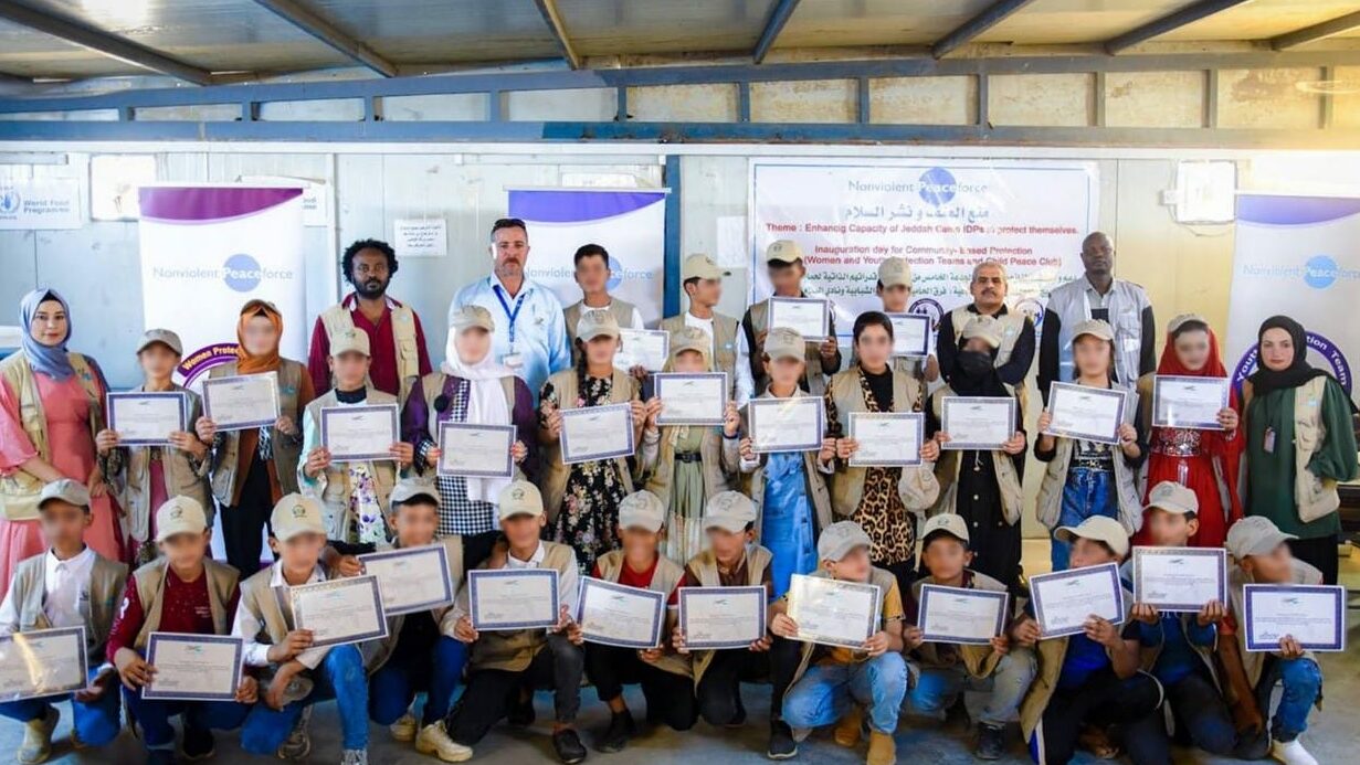 A group of young people holding certificates poses with adults in front of a building. The group is diverse in age and dress. There are banners on the wall behind them. The setting appears to be an award or graduation event.