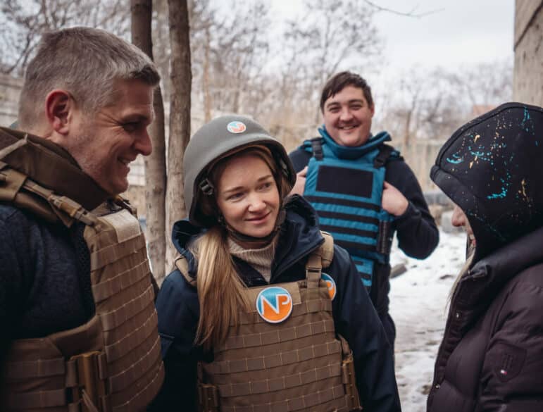 Four people wearing protective vests and winter clothing stand outdoors in a snowy area. One person is wearing a helmet; others are smiling or looking at each other. Trees and a concrete building are visible in the background.