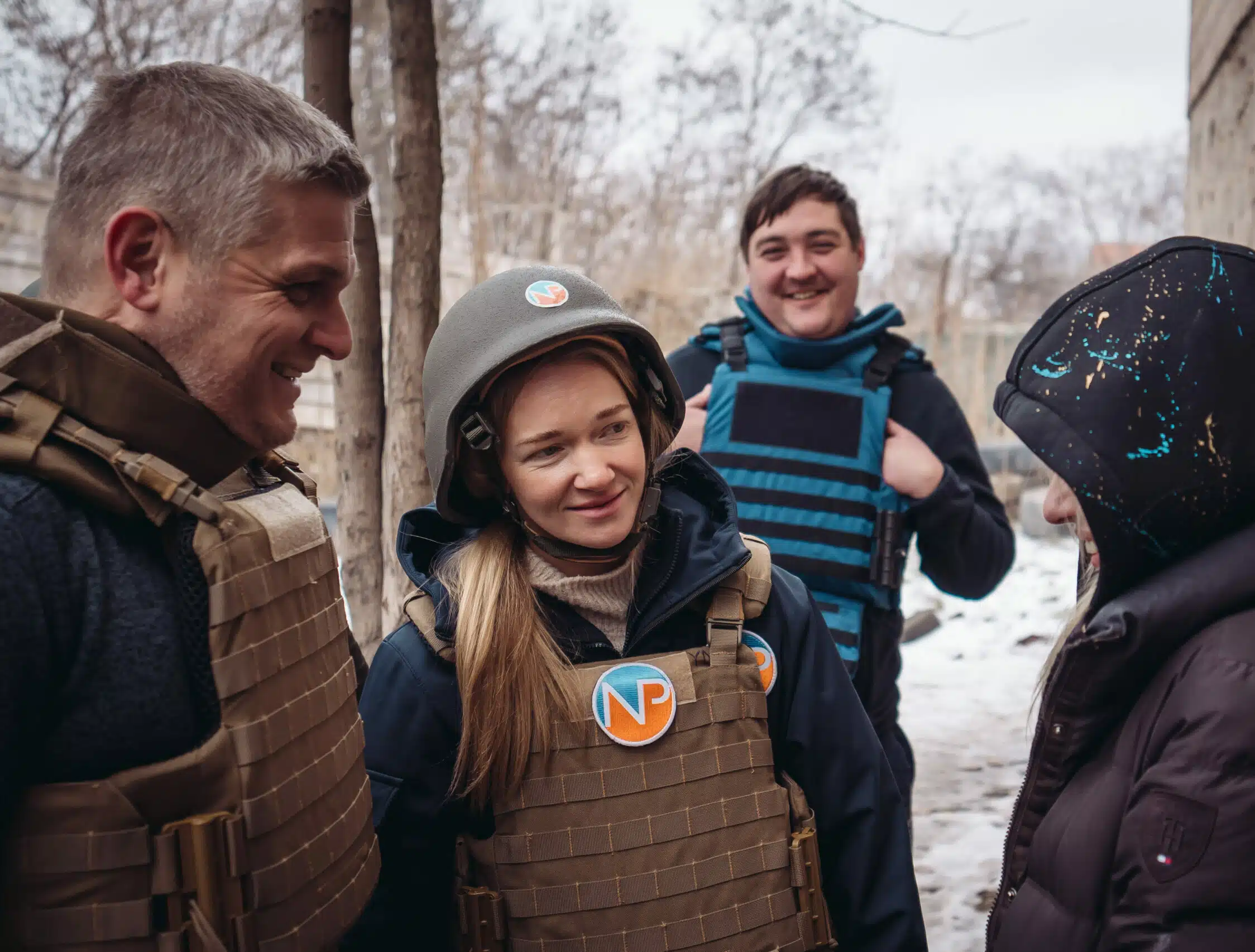 Four people wearing protective vests and winter clothing stand outdoors in a snowy area. One person is wearing a helmet; others are smiling or looking at each other. Trees and a concrete building are visible in the background.