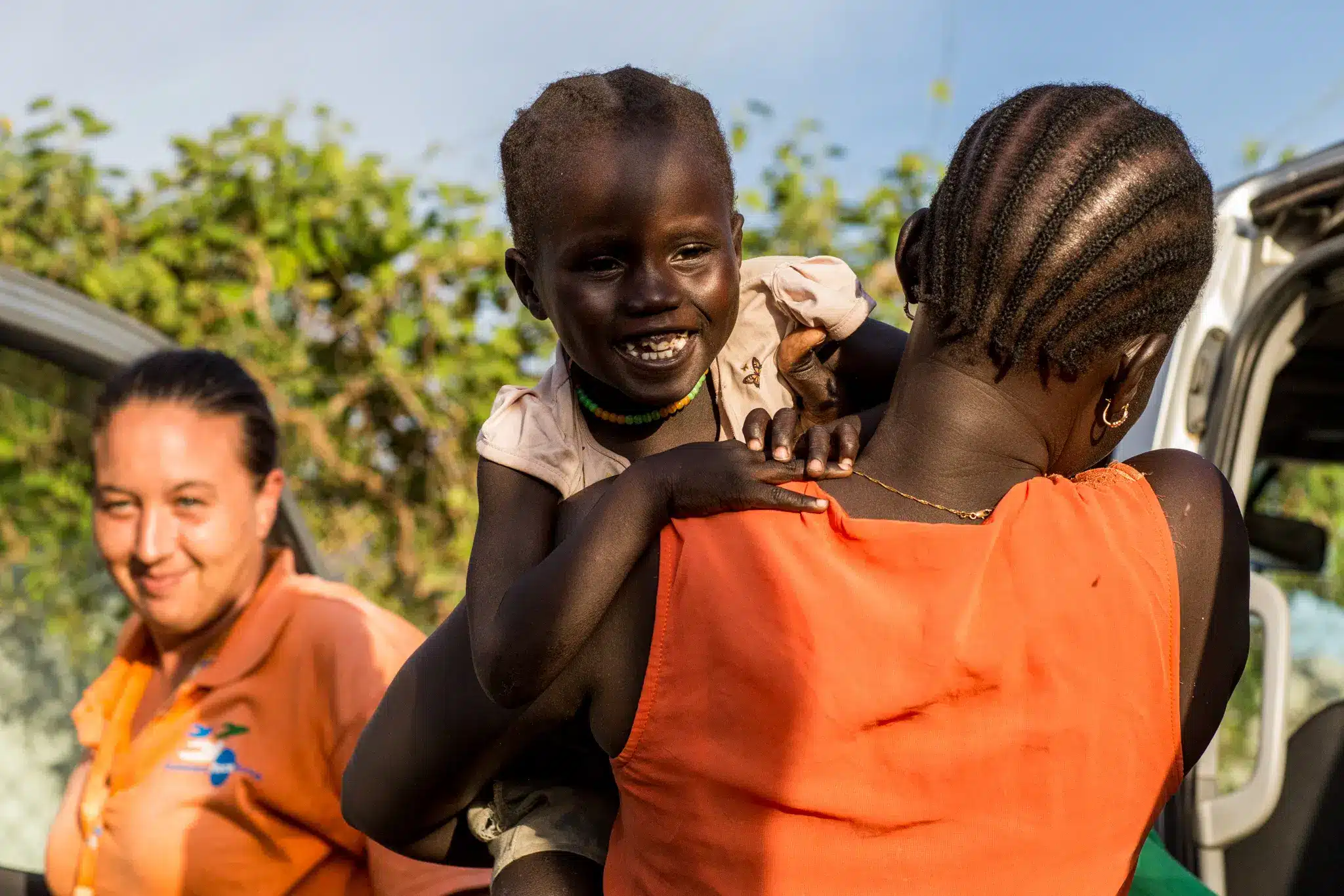 A woman in an orange dress holds a smiling child on her shoulder, while another woman in an orange shirt stands nearby. There is greenery and part of a vehicle in the background.