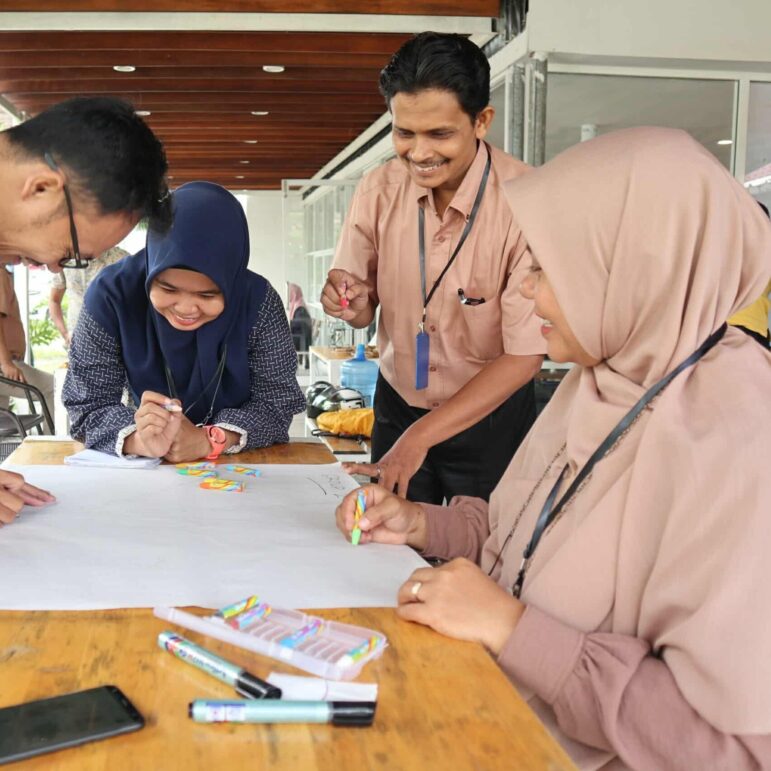 Four adults sit and stand around a wooden table, drawing and writing on a large sheet of paper with colored markers. They appear to be collaborating in a casual indoor setting.