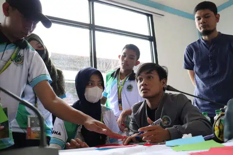 Fünf Personen sitzen um einen Tisch mit Papier, Stiften und Notizbüchern. Eine Person deutet auf Dokumente, während die anderen zusehen oder zuhören. Eine Frau trägt Maske und Hidschab. Im Hintergrund sind große Fenster zu sehen.