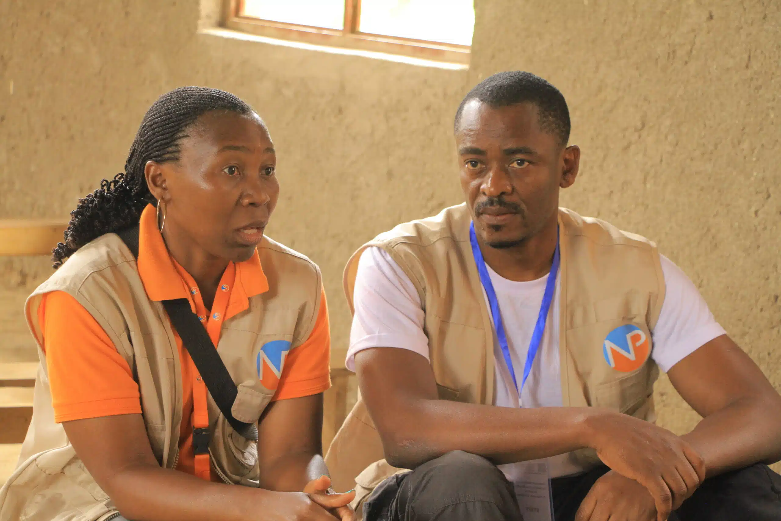 Two people in tan vests with blue and orange logos sit indoors against a bare wall, appearing to listen attentively. A window is visible in the background.