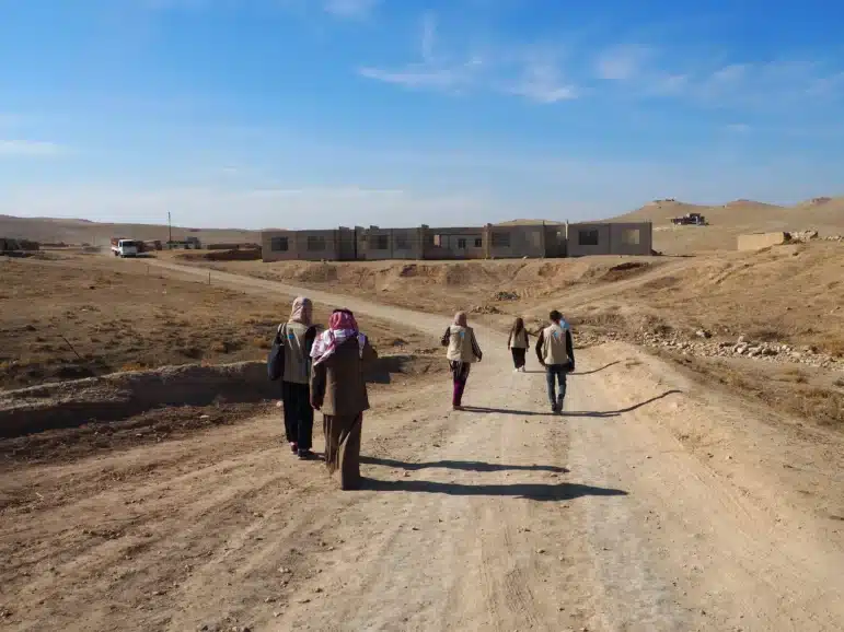 Five people walk on a dirt road toward a group of low, unfinished buildings in a dry, barren landscape under a blue sky with scattered clouds. Some wear headscarves and carry bags or backpacks.