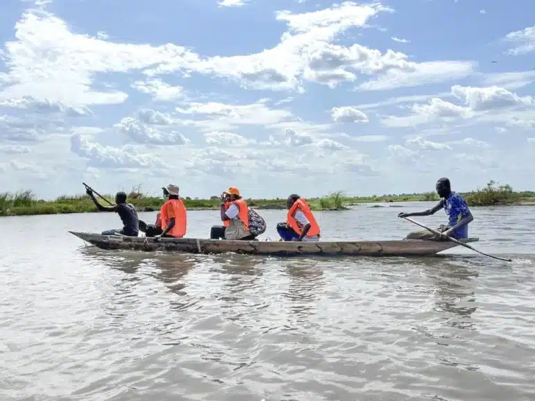 Five people in a wooden canoe on a river, four wearing orange life jackets, with one person paddling at the back. The sky is partly cloudy, and the water reflects the scenery.