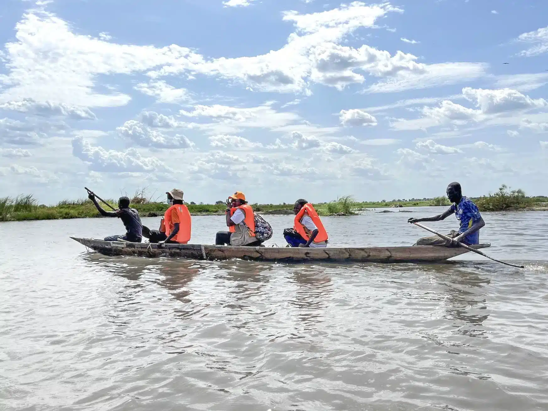 Five people in a wooden canoe on a river, four wearing orange life jackets, with one person paddling at the back. The sky is partly cloudy, and the water reflects the scenery.
