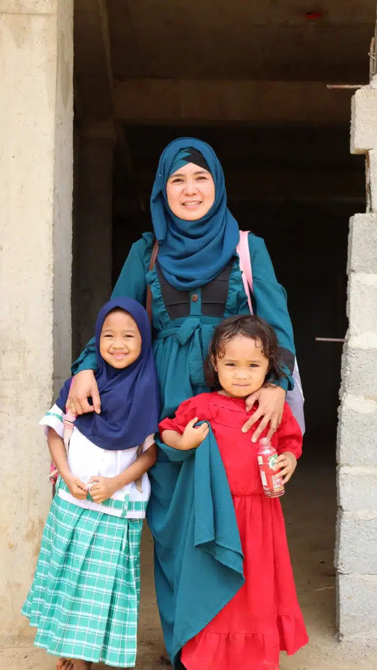 Una mujer con un vestido turquesa y un hiyab está de pie junto a dos niñas pequeñas, una con un hiyab azul y una falda a cuadros, y la otra con un vestido rojo, frente a un edificio de hormigón en construcción.