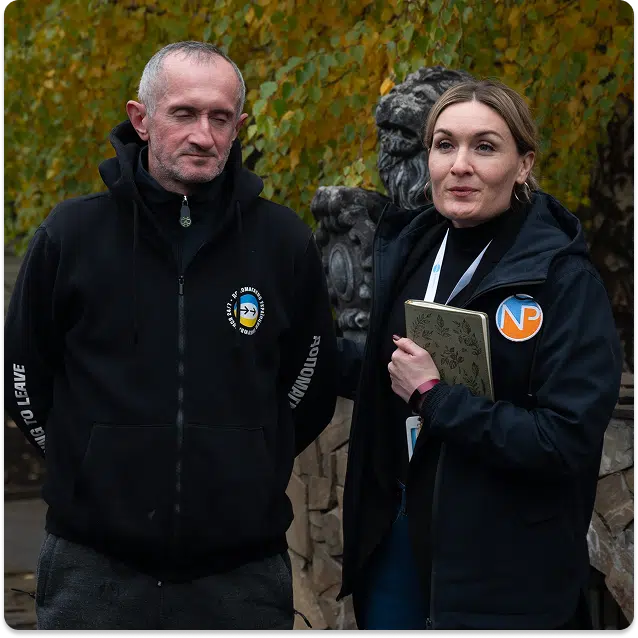 Two people stand outdoors in front of stone sculptures and autumn trees. Both wear black jackets with logos; the man looks ahead with hands clasped, while the woman holds a notebook and appears to be speaking.