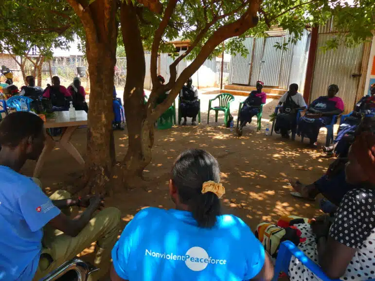 A group of people sit in chairs arranged in a circle under a large tree for a meeting, with one participant wearing a blue shirt that reads NonviolentPeaceforce.