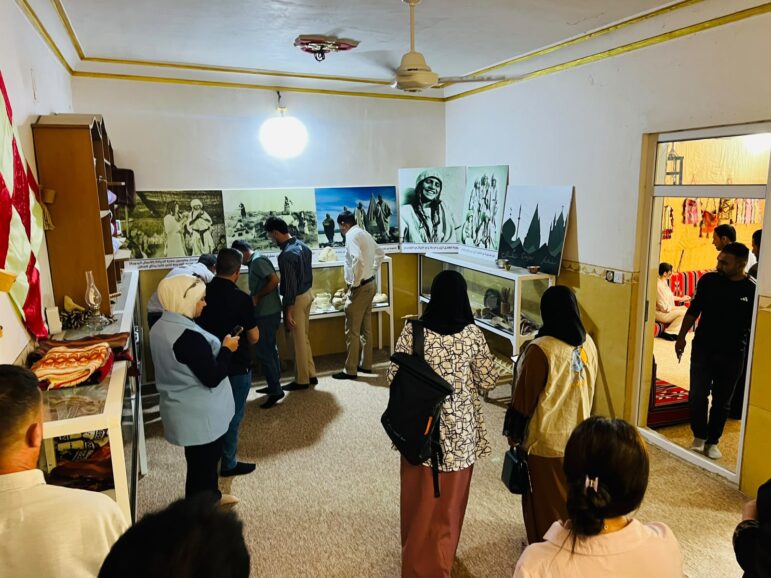 A group of people viewing historical photographs and artifacts displayed in an indoor exhibition space.